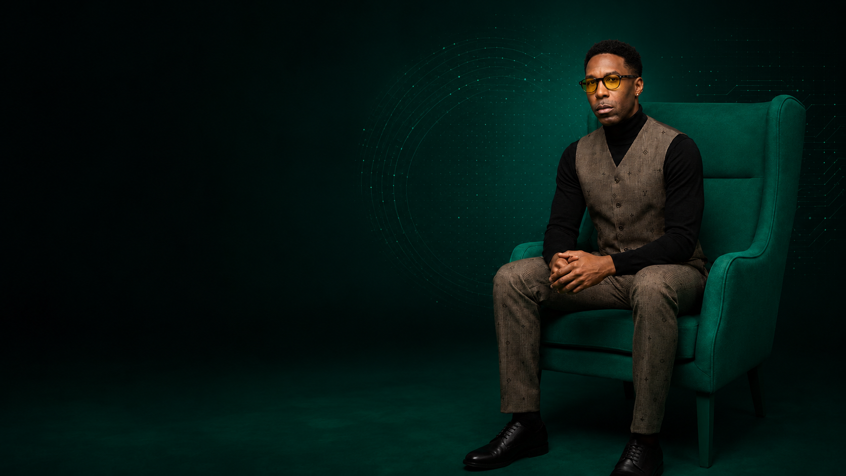 Lajuane Torrey seated in a green chair wearing a brown vest, black turtleneck, glasses, and dress shoes.